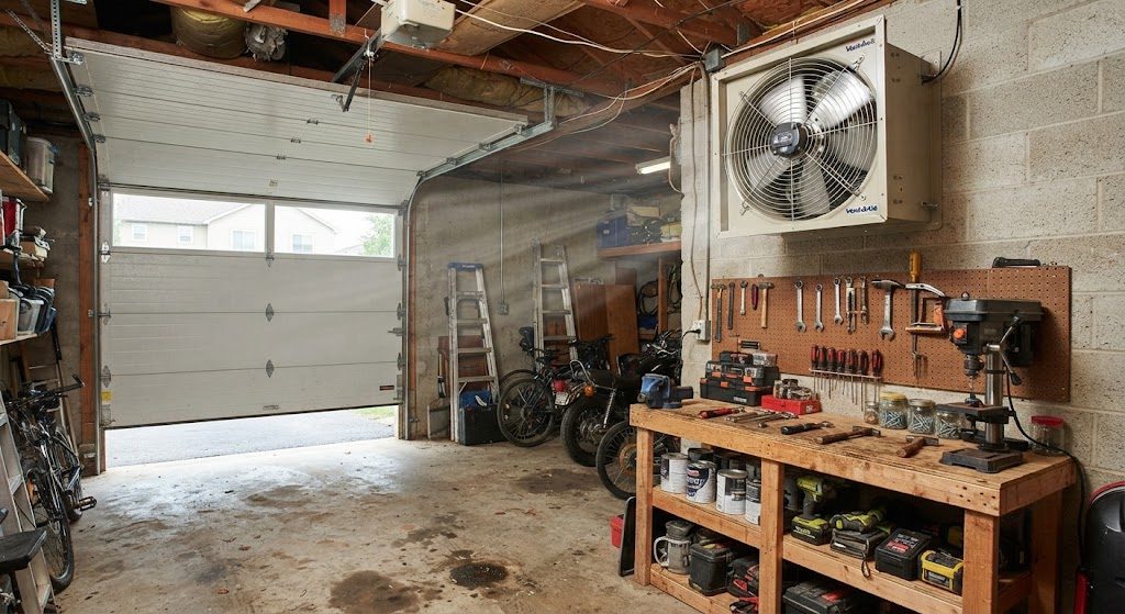 Wall-mounted exhaust fan installed near the ceiling of a garage with the door cracked open for cross-flow ventilation
