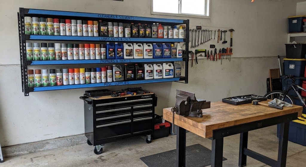 Steel shelving unit in a garage with labeled shelves for fluids, spray cans, and parts, a rolling tool cart parked below